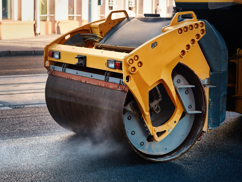 A steamroller with a large yellow metal drum, under the expertise of a commercial asphalt company in Osceola County FL, compacts fresh asphalt on a road.