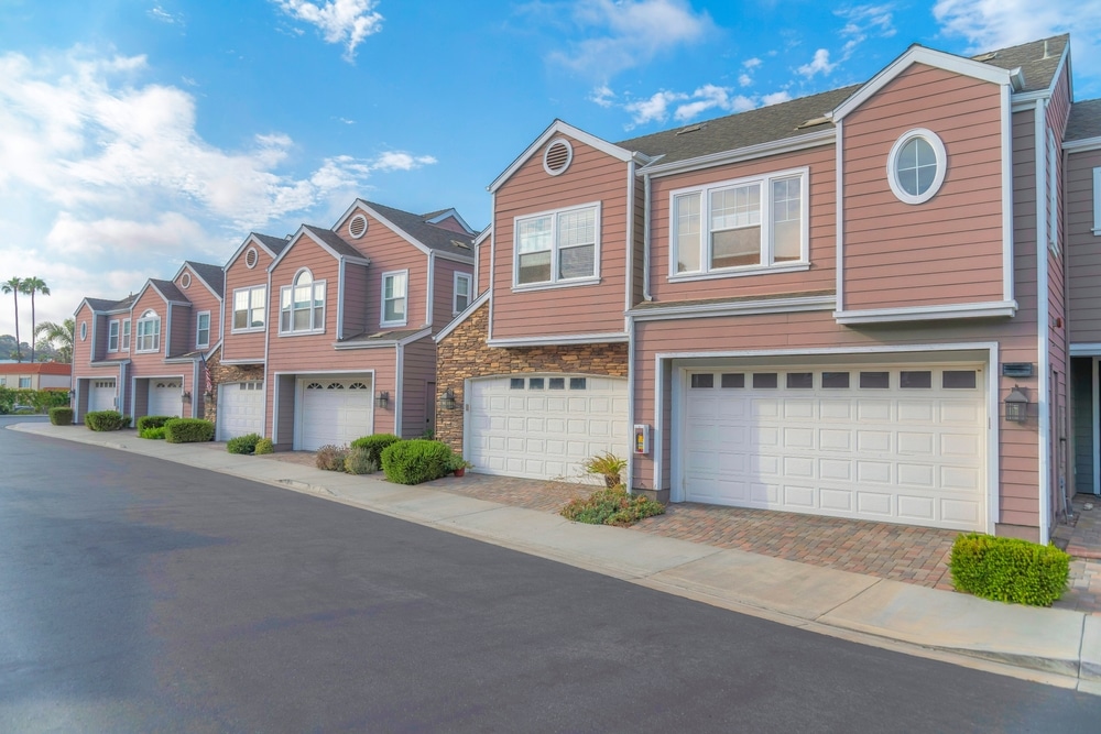 A row of modern, two-story townhouses with pink siding and white trim. Each unit features a garage and a small front yard with greenery. The street, newly paved by a commercial asphalt company in Osceola County FL, is empty under a sky with scattered clouds.