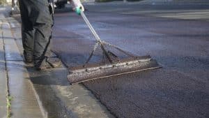 A worker from a commercial asphalt company in Osceola County, FL, uses a large metal tool to spread asphalt on a road surface. Dressed in protective clothing, including boots and gloves, the close-up scene shows the road partially covered in newly laid asphalt.