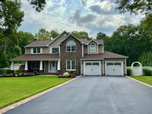 A two-story house with a stone and siding exterior, featuring a manicured lawn, flower beds, and an expertly paved long driveway leading to a two-car garage. Tall trees surround the property under partly cloudy skies.