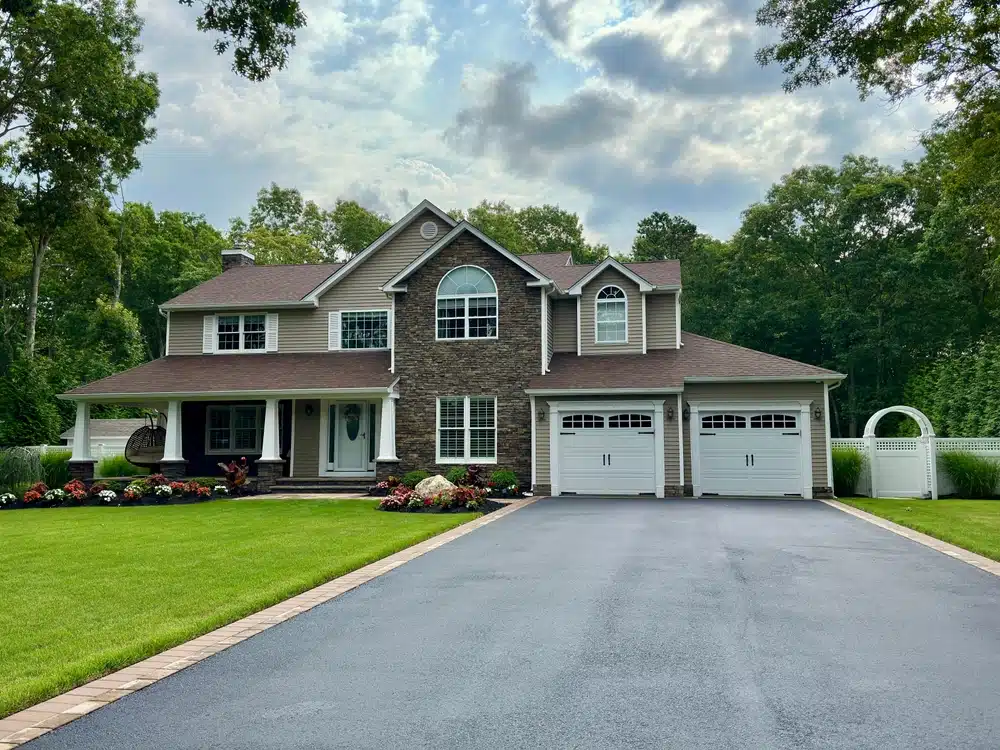 A two-story house with a stone and siding exterior, featuring a manicured lawn, flower beds, and an expertly paved long driveway leading to a two-car garage. Tall trees surround the property under partly cloudy skies.