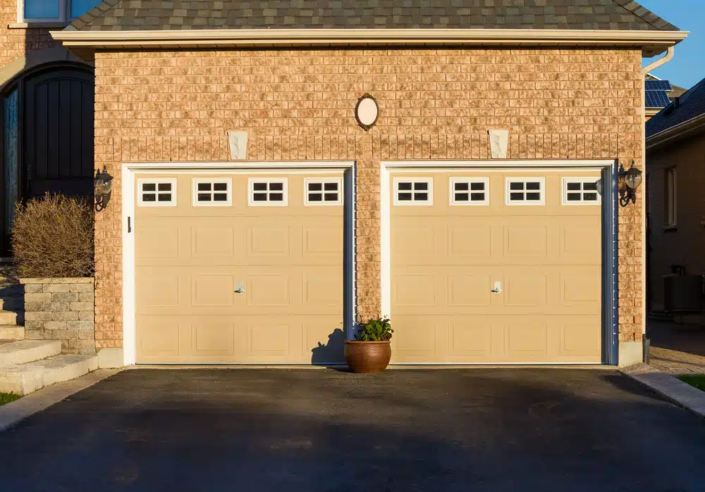 A suburban home with two beige garage doors featuring small square windows at the top sits gracefully. Above the garages are wall-mounted lanterns against a brick exterior, reminiscent of work by a commercial paving company. A central light fixture and potted plant add charm to the driveway.